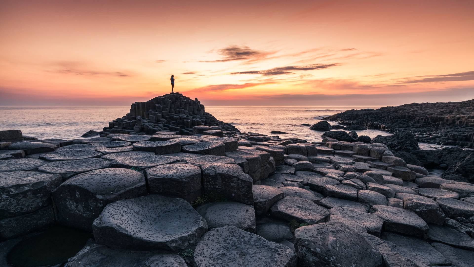 A person stands atop the hexagonal basalt columns of Giant’s Causeway in Northern Ireland at sunset, with a colorful sky and calm sea in the background.