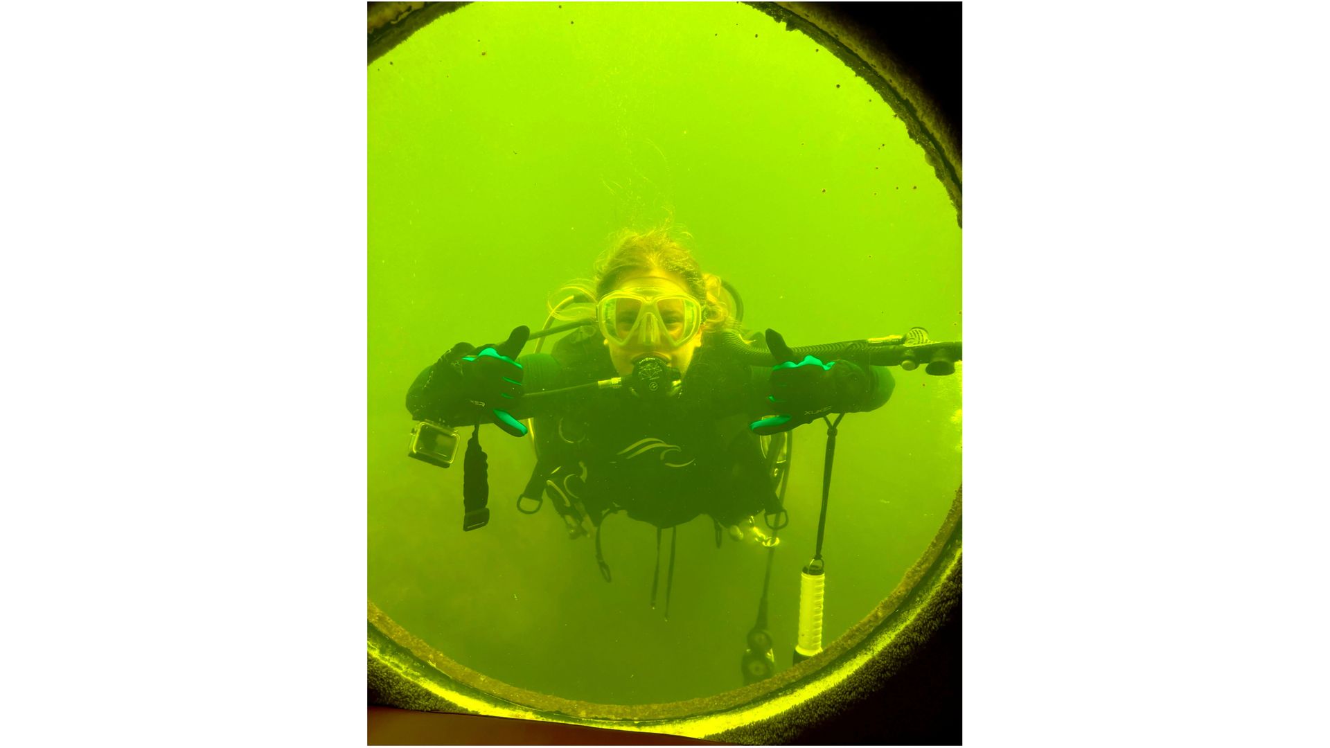 photo through a porthole showing a woman scuba diving in greenish-tinted water