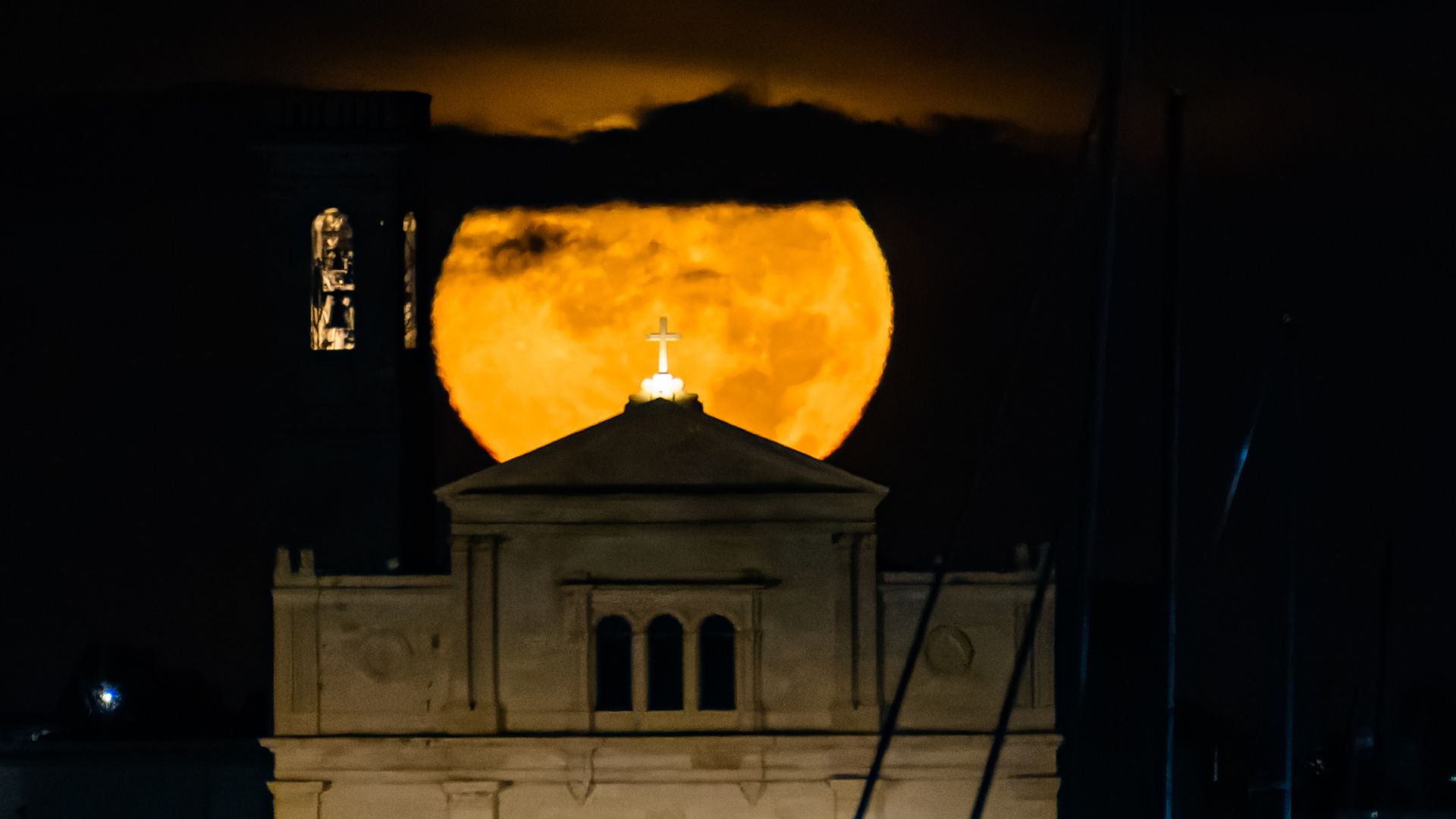 An orange full moon shines behind a church at night as clouds streak its surface.