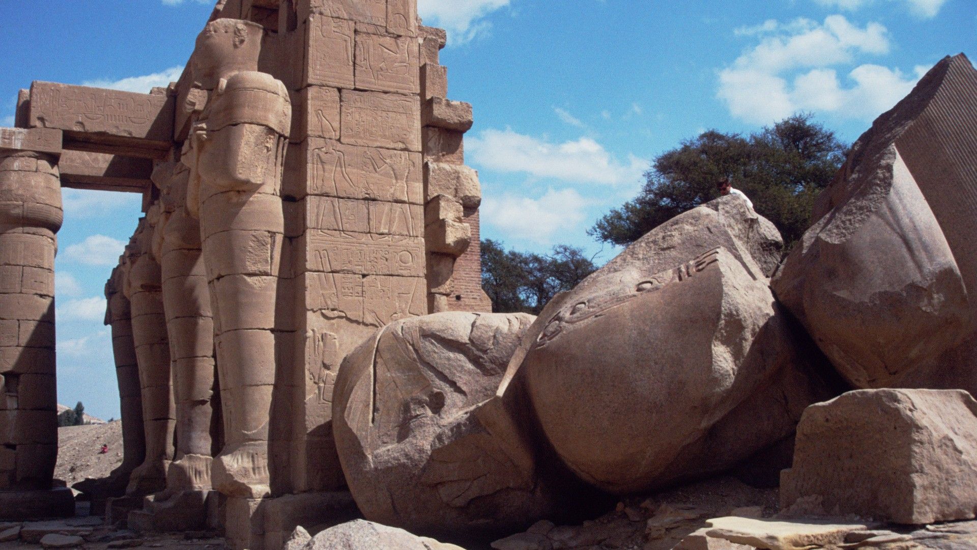 Sunlit ancient Egyptian temple ruins, with towering carved columns and human statues. AT the forefront is a giant fallen statue broken to pieces.