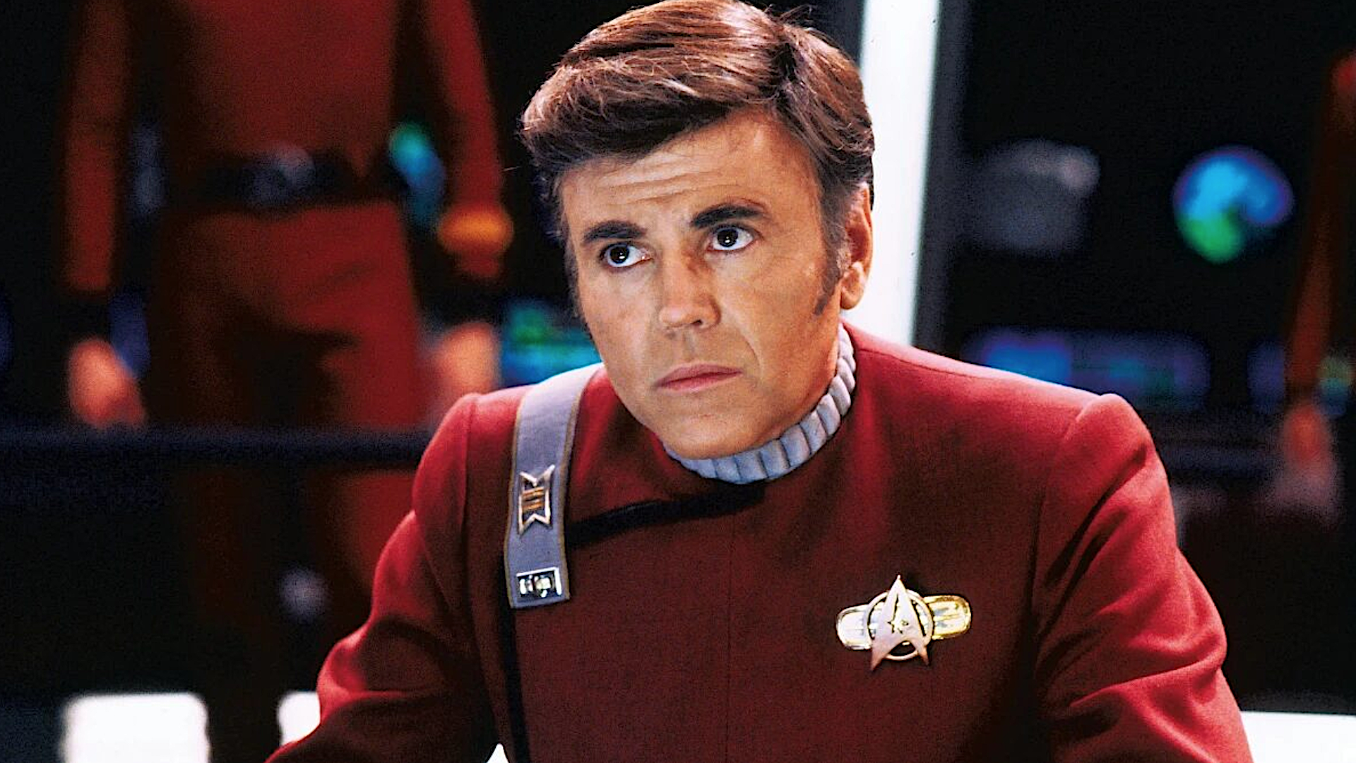 a man at a starship bridge in a red uniform