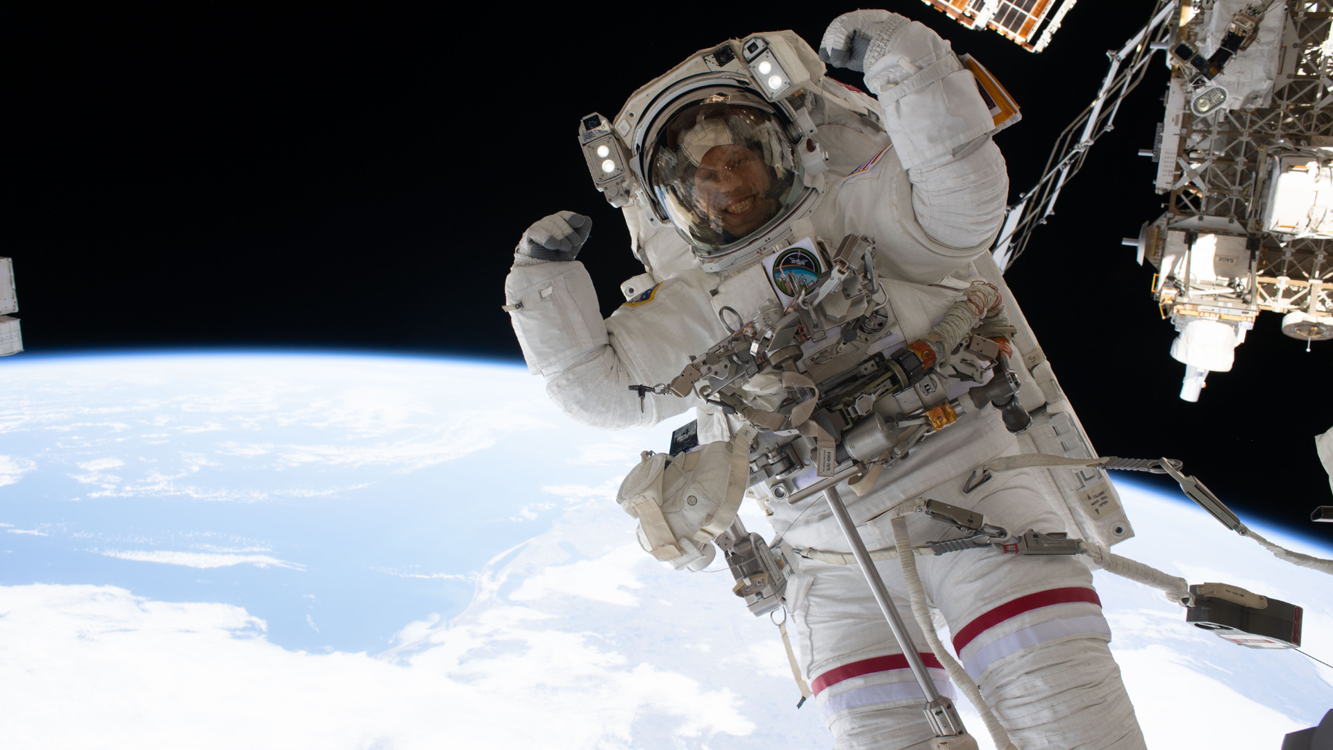 an astronaut in a white spacesuit flexes on a spacewalk, with earth in the background