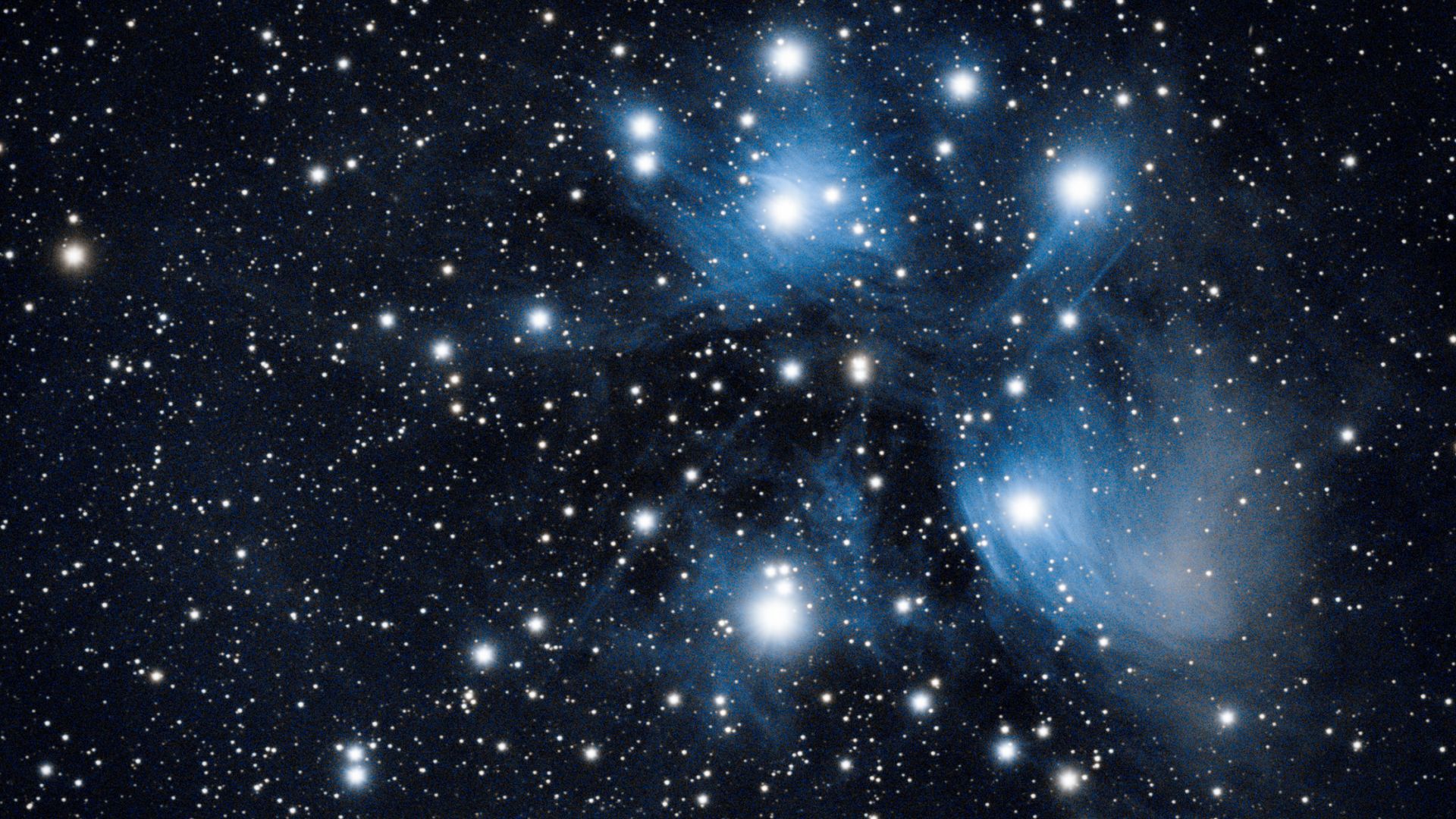 A collection of blue-white stars glow against a field of dimmer stars in the blackness of space. A faint nebulosity can be seen glowing before the brightest stars.