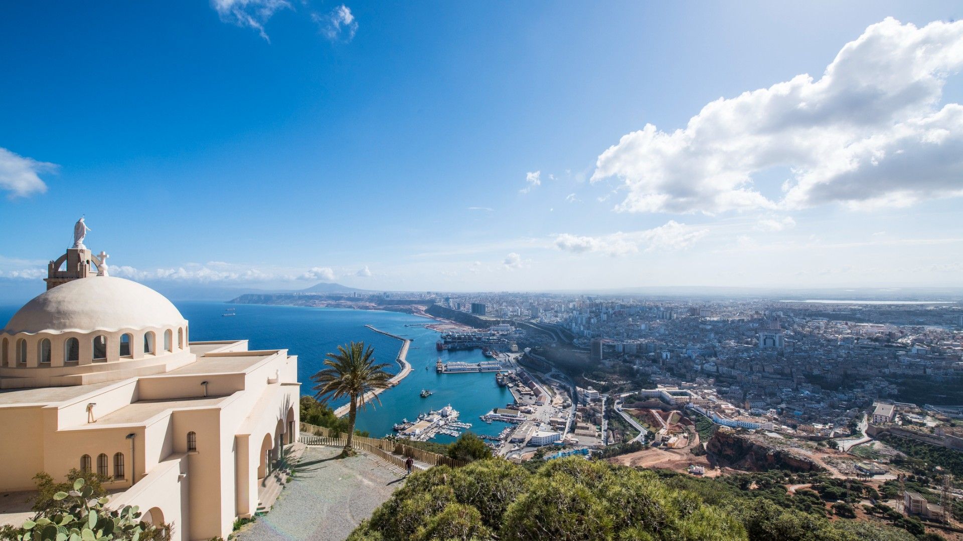 Sunlit hillside panorama of a cream-colored domed church overlooking a blue bay, marina, and sprawling coastal city beneath a bright, cloud-dotted sky.