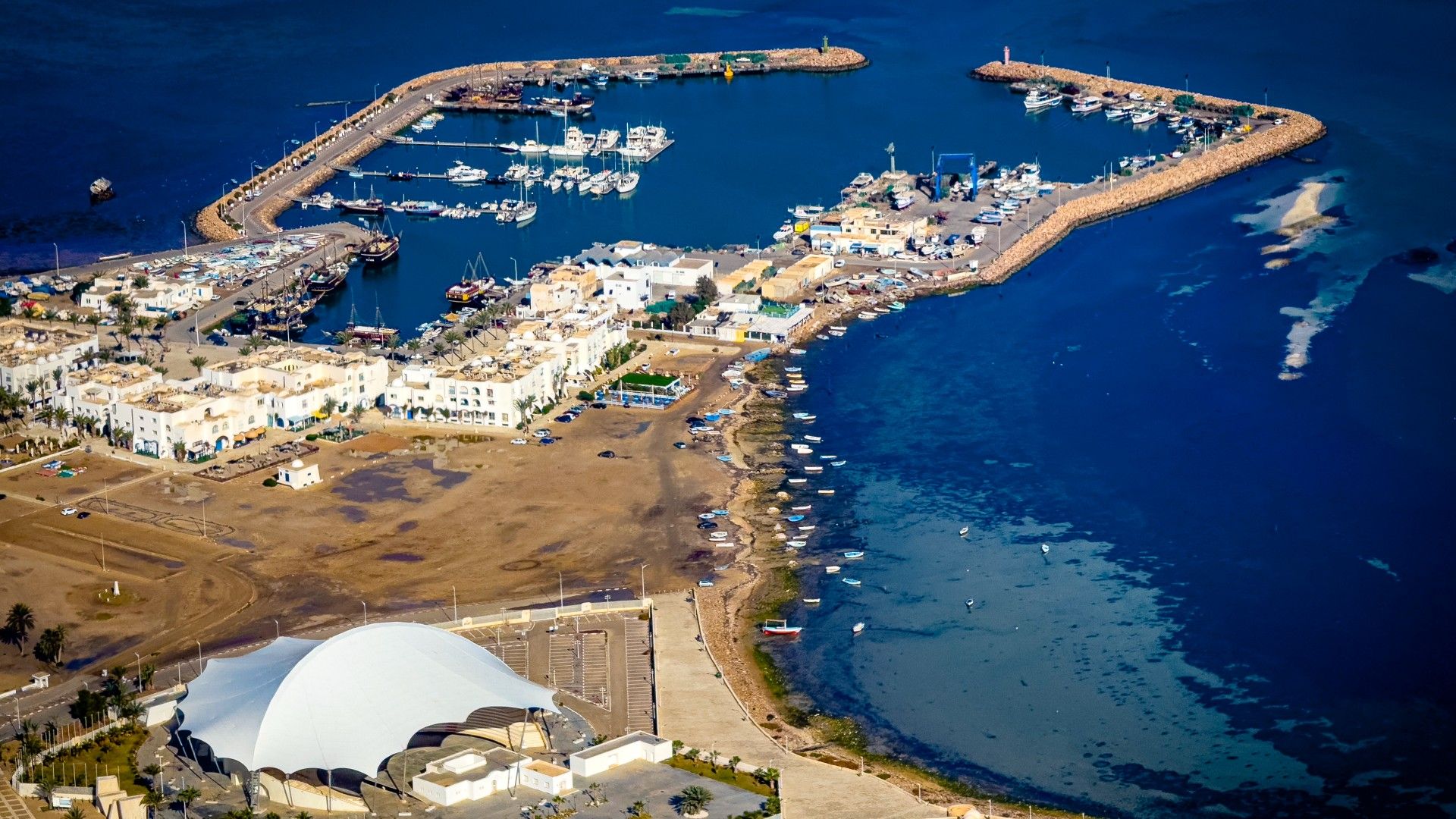 Aerial view of a coastal town featuring a marina with docked boats, a large white amphitheater, and an ancient stone fortress near the water.