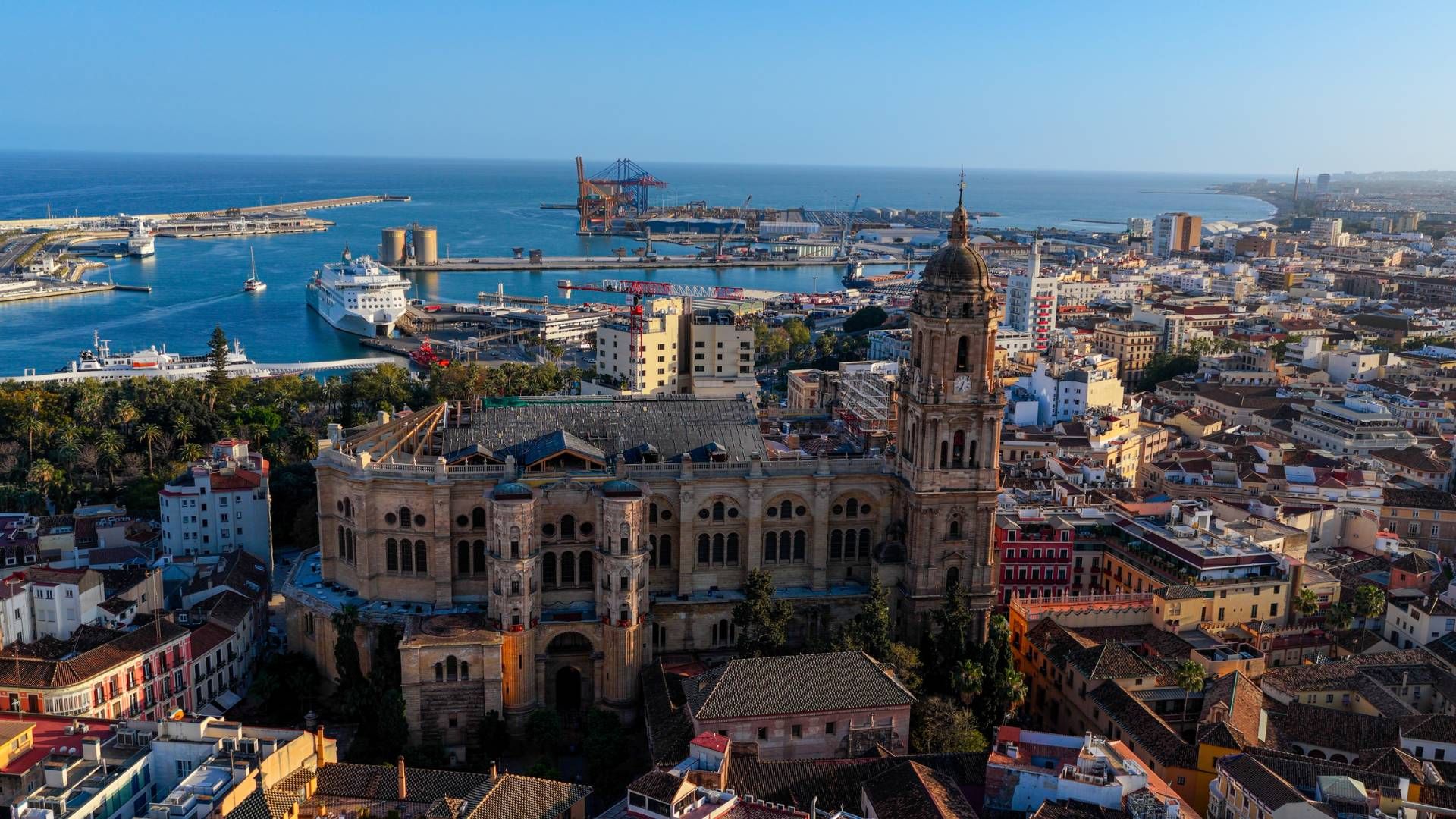 Aerial view of the giant Málaga Cathedral and historic dense city center at sunset. In the distance you can see the palm tree-lined harbour.