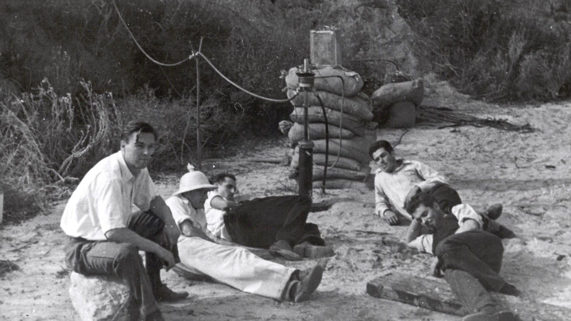 five men in white button-down shirts lay on the desert sand next to sandbags and wires held up on pylons