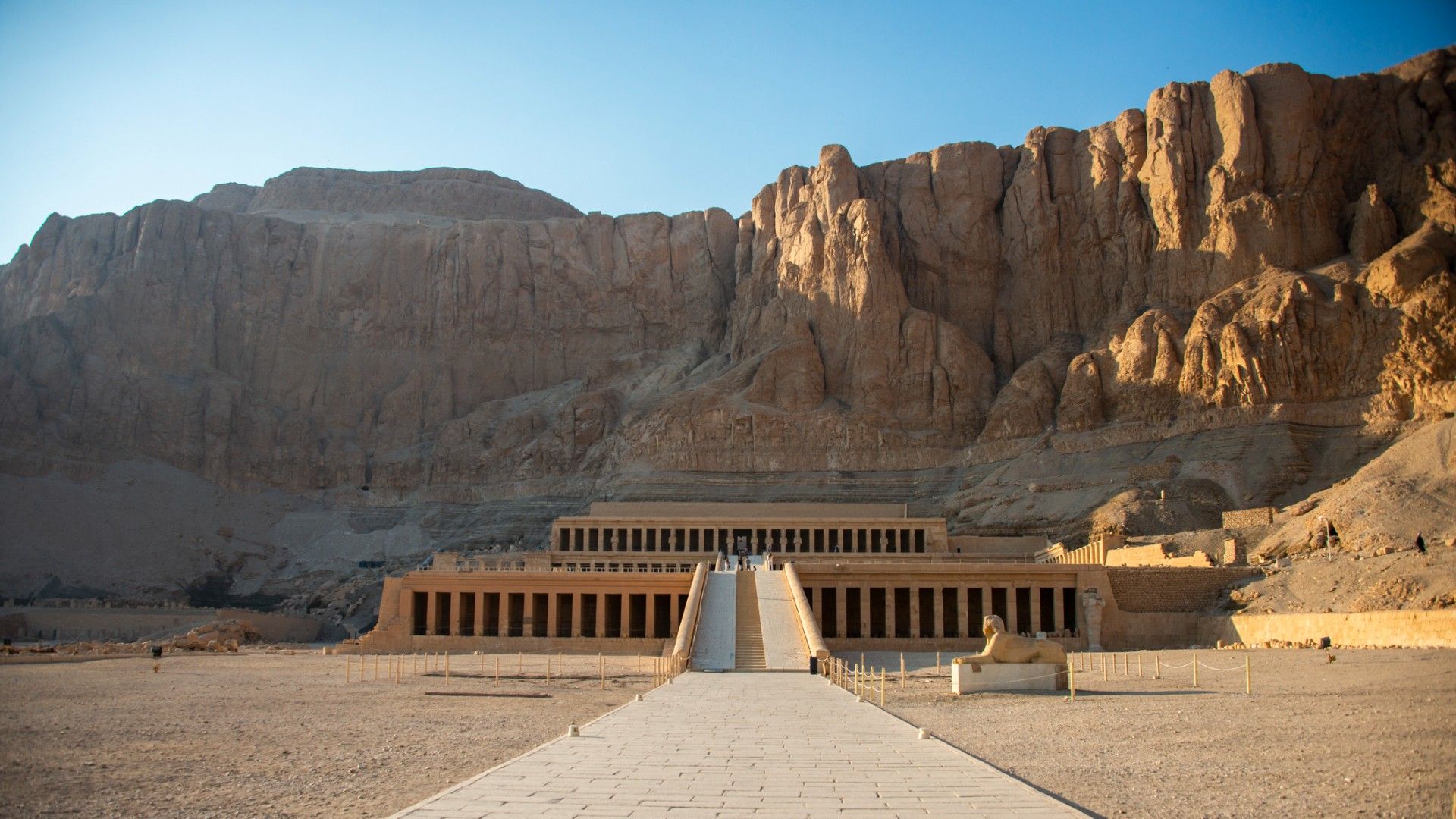 Hatshepsut’s ancient Egyptian temple stands at the end of a long stone walkway beneath towering desert cliffs, evoking a quiet, sunlit grandeur.