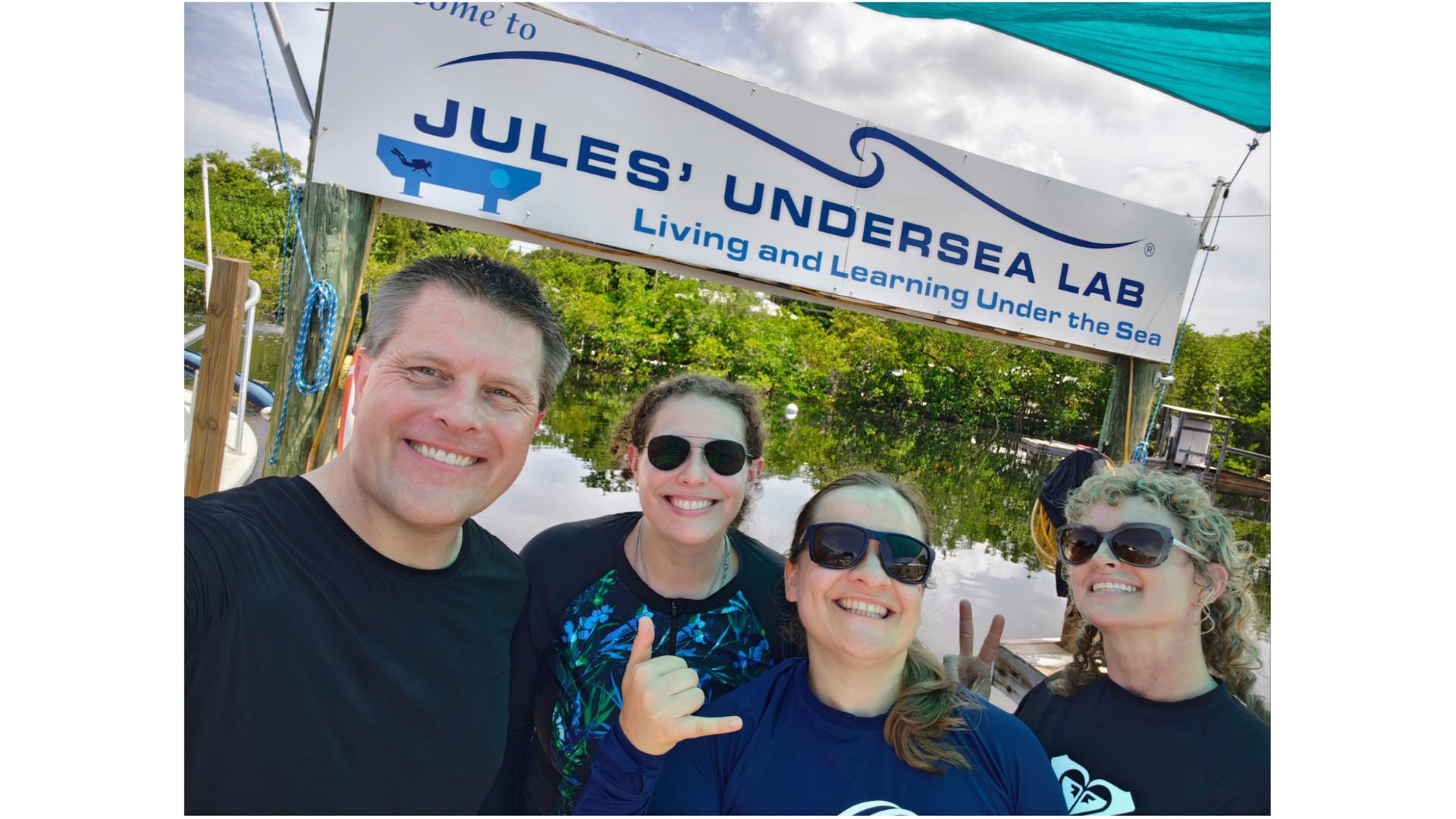 photo of four smiling people, three women and one man, standing outside by a sign that reads 