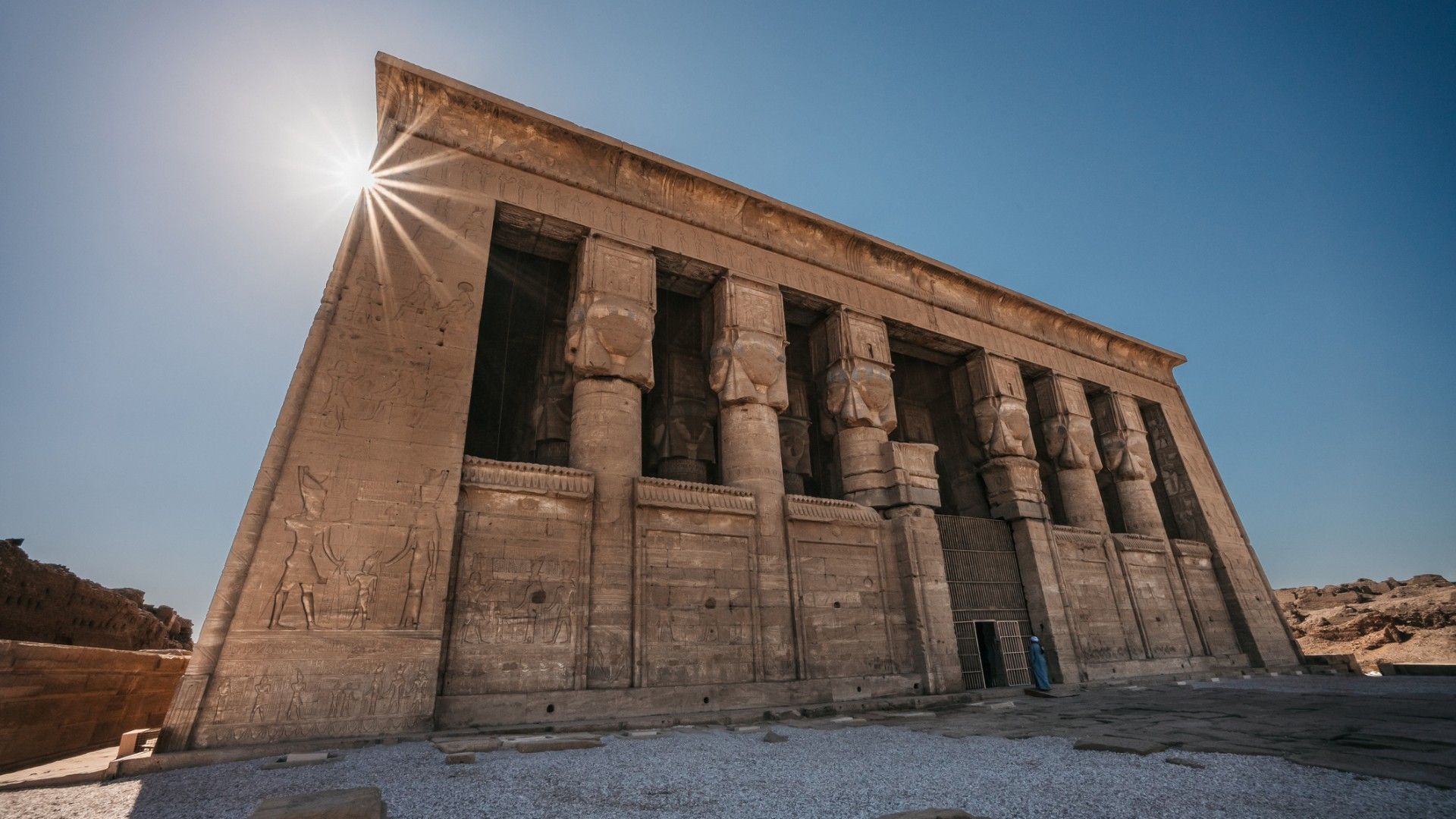 Sunlight flares over an ancient Egyptian sandstone temple, its carved reliefs and towering columns rising against a clear blue sky in the desert.