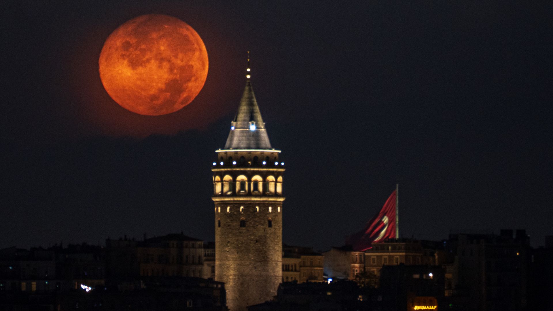 A red full moon is photographed shining next to an illuminated medieval tower at night.